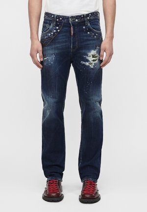 Mand iført mørkeblå slidte jeans med metaløjer og en camouflagelap på højre lår, kombineret med brune støvler og røde snørebånd.