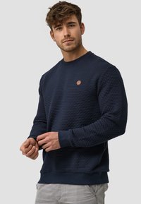 Marineblauwe sweatshirt met een gestructureerd honingraatpatroon, geribde mouwen en zoom; voorzien van een klein bruin logopatch op de borst.