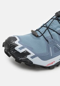 Chaussure de trail running en bleu clair, avec un dessus texturé, un embout en caoutchouc noir orné d'un motif angulaire blanc, et un système de laçage sécurisé.