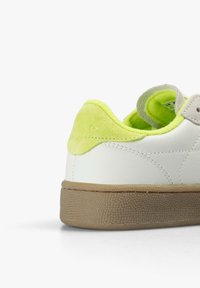 Baskets blanches avec talon en daim jaune fluo et doublure, semelle texturée marron clair, et couture visible sur le côté.