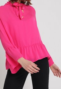 Blusa rosa brillante realizzata in tessuto leggero, con scollo a legatura, un'apertura a chiave e un orlo peplum a sbalzo con maniche lunghe.