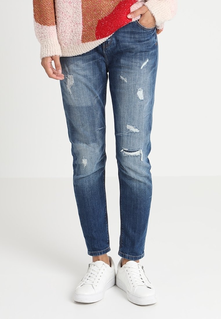 Vybledlé modré džíny s opotřebovanými detaily, zdobené nášivkami a třepenými okraji, kombinované s bílými teniskami, vystavené na neutrálním pozadí.