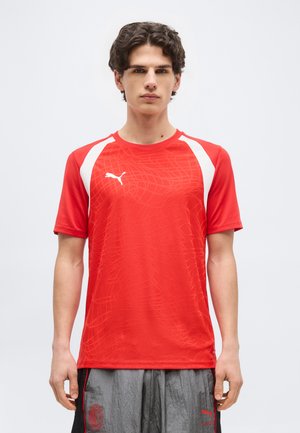 Młody mężczyzna stoi przodem, ubrany w czerwoną sportową koszulkę Puma z białymi akcentami oraz szare spodenki Puma.