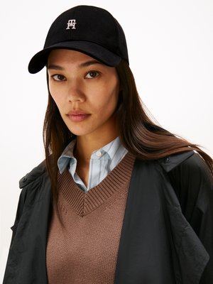 Cappello nero con logo, tessuto testurizzato, indossato con una camicia azzurra a collo e un maglione lavorato a maglia marrone, stratificato con una giacca nera.
