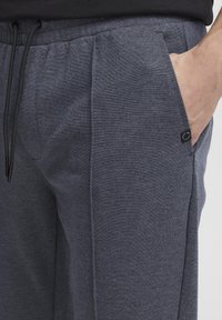 Un pantalon de jogging gris en tissu texturé avec un waistband élastique et cordon de serrage, ainsi que des poches latérales. Une petite étiquette de logo est visible sur la poche.