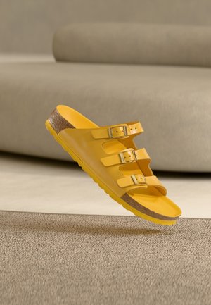 Gele sandaal met drie verstelbare gespen en kurkzool die zweeft boven een beige, gestructureerd tapijt in een kamer met neutrale tinten.