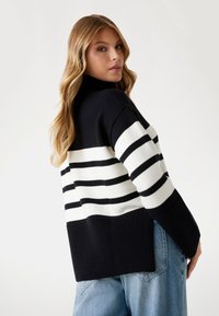 Maglione a righe bianco e nero in maglia con collo alto, polsi e orlo a costine; vestibilità ampia, con una larga banda di righe al centro.