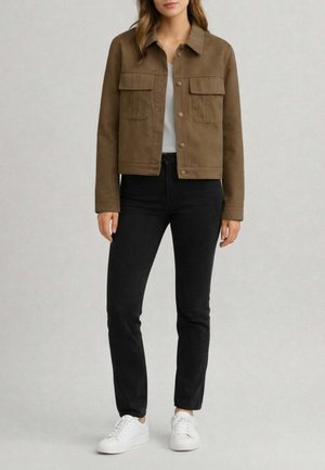 Donna che indossa una giacca marrone con bottoni e tasche a patta, jeans neri, sneakers bianche e una maglietta bianca, su uno sfondo chiaro e uniforme.