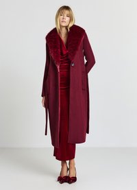 Langer burgunderroter Mantel mit Kunstpelzkragen, mit Knopfleiste und zwei Seitentaschen, getragen über einem passenden Samtkleid und Schuhen mit Absatz.