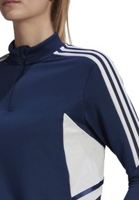 Marineblauw sporttop met een hoge kraag, voorzien van witte zijpanelen en drie witte strepen langs de schouders. Gemaakt van een gladde, rekbare stof.