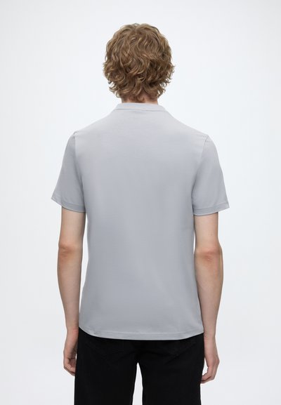 Jeune personne aux cheveux bouclés portant un t-shirt uni gris clair à manches courtes et un pantalon noir, vue de dos sur fond blanc.