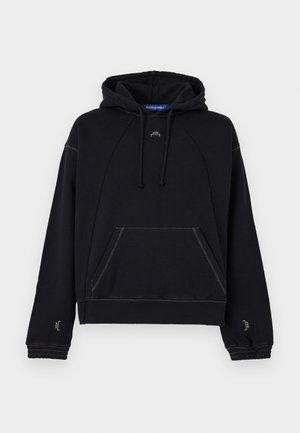 Hombre con cabello rizado que lleva una sudadera negra con bolsillo delantero y pantalones cargo grises, de pie frente a un fondo claro y liso.