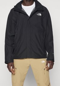 Černá voděodolná bunda se zipem vpředu a vysokým límcem. Na hrudi se nachází logo a má elastické manžety. Doplněna béžovými kalhotami.