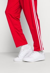 Röda sportbyxor med vita ränder på sidorna, kombinerat med vita sneakers. Sneakers har en slät lädertextur och snördesign.