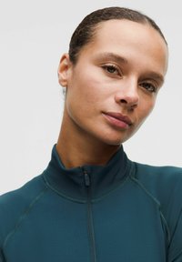 Zbliżenie na kobietę z włosami związanymi w kok, ubraną w ciemnoniebieską, zapinaną na zamek kurtkę, spokojnie patrzącą w kamerę na jednolitym tle.