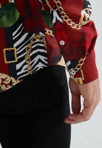 Chemise rouge à motifs comprenant des roses, des chaînes et des rayures zèbres ; tissu lisse ; poignet à boutons avec un bouton blanc ; jeans noirs partiellement visibles.
