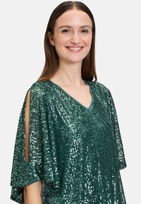 Top verde con paillettes, scollatura a V, maniche corte e aperture sulle spalle. Presenta una texture scintillante e un design morbido e fluido.