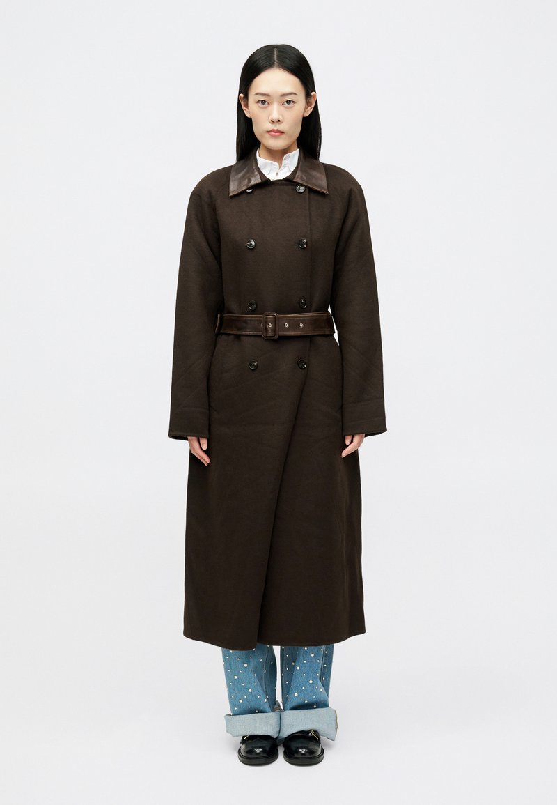 Καφέ διπλόχρωμο παλτό με δερμάτινο γιακά και ταιριαστό ζωνάκι, συνδυασμένο με ανοιχτόχρωμα τζιν παντελόνια με λευκές βούλες.