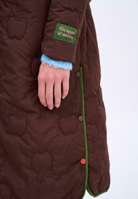 Brązowy pikowany płaszcz z motywem jabłka, zielonym wykończeniem i kolorowymi guzikami. Widoczna ręka nosi niebieską, puszystą rękawiczkę pod płaszczem.