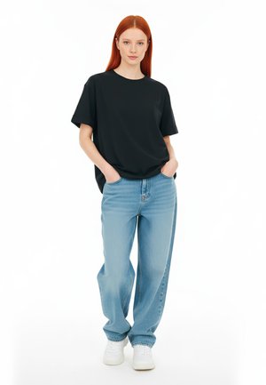 Mlada ženska z dolgimi rdečimi lasmi, oblečena v črn majico, ohlapne svetlo modre jeans hlače in bele športne copate, stoji s rokama v žepih.