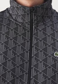 Gros plan d'une veste noire et grise à motif géométrique avec un petit logo crocodile vert sur le côté droit.