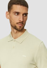 Khaki polo z teksturowanej tkaniny, z kołnierzykiem, dwoma guzikami i małym białym logo na lewej piersi.