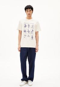 Lichtbeige T-shirt met natuurthema in blauwe illustraties en de tekst "BEWAAR DE NATUUR", gecombineerd met donkerblauwe losse broek en witte sneakers.