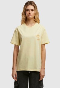 Gelbes Baumwoll-T-Shirt mit kurzen Ärmeln, das ein kleines oranges Charakter-Graphic im linken Brustbereich zeigt. Schlichte und entspannte Passform.