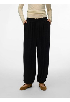 La personne porte un pantalon noir ample avec des poignets élastiques, un haut beige à manches longues, les mains dans les poches, et des chaussures plates marron avec des brides.