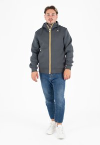 Grijze hoodie met rits, voorzien van gele en witte gestreepte accenten, voorzakken en een verstelbare capuchon. Gepaard met blauwe spijkerbroek en witte schoenen.