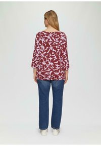 Bordeaux top met lichtpaarse bloemenprint, 3/4 mouwen, losse pasvorm. Gecombineerd met blauwe jeans en witte sneakers, gezien vanaf de achterkant.