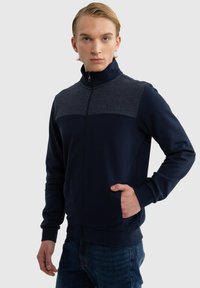 Mornarska zip majica s kontrastnim sivim panelom na vrhu, rebrastim manžetama i bočnom džepom. Glatka tekstura tkanine.