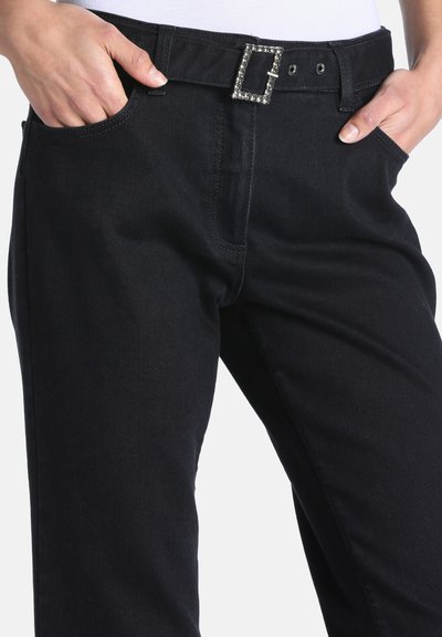 Mains dans les poches avant d’un pantalon noir avec une boucle de ceinture rectangulaire ornée de strass, portée avec un haut blanc.
