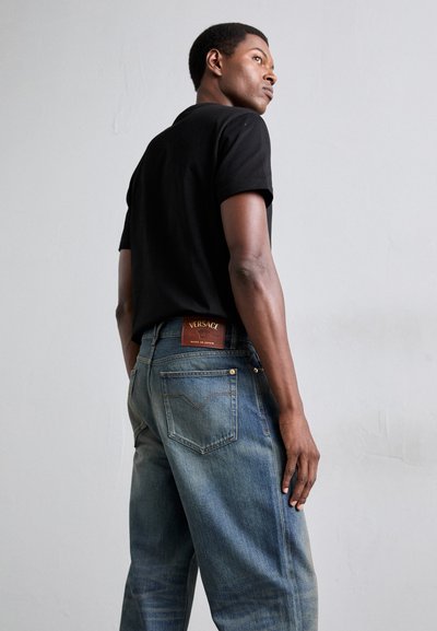 Polo shirt negru cu mânecă scurtă asociat cu blugi bleu cu talie medie, având o etichetă din piele și o textură de denim cu efecte de uzură și detalii de cusătură.