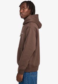 Brauner Kapuzenpullover mit einer Fronttasche, aus weichem Stoff gefertigt. Verfügt über Kordelzüge und gerippte Bündchen, kombiniert mit Jeans.