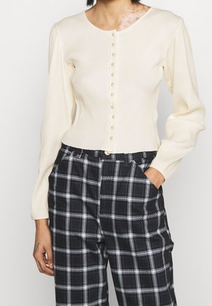 Persoon draagt een crème geribbelde vest met knopen en zwart-witte geruite broek, één hand in de zak, tegen een effen achtergrond.