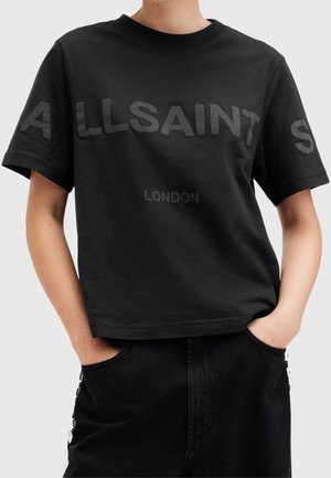Persoon draagt een zwart cropped T-shirt met de tekst "ALLSAINTS LONDON" op de borst, handen in de zakken, gecombineerd met een zwarte broek.