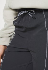 Zwarte sportbroek met een verstelbare tailleband, voorzien van contrasterende witte stiksels en een gladde textuur. Close-up van de taildetails.