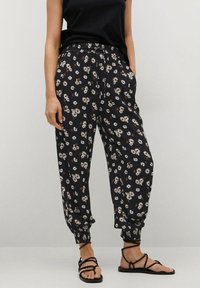 Femme portant un pantalon noir ample avec des motifs de marguerites blanches, un haut sans manches noir, et des sandales plates noires à lanières, se tenant avec les mains dans les poches.