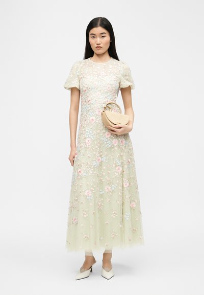 Γυναίκα που φοράει μεσαίο φόρεμα σε απαλό πράσινο με κεντήματα λουλουδιών και φουσκωτά μανίκια, κρατώντας μια μικρή μπεζ τσάντα και φορόντας λευκές μυτερές γόβες.