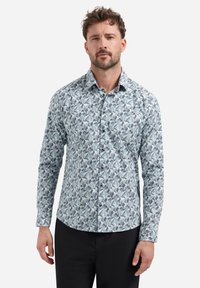 Man in een langarmlijk overhemd met een blauw-grijze abstracte geometrische patroon en zwarte broek, staand tegen een witte achtergrond.