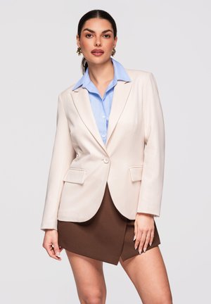 Žena oblečená v krémovém saku přes modrou košili s límečkem a hnědou asymetrickou sukni, stojící před jednoduchým světlým pozadím.