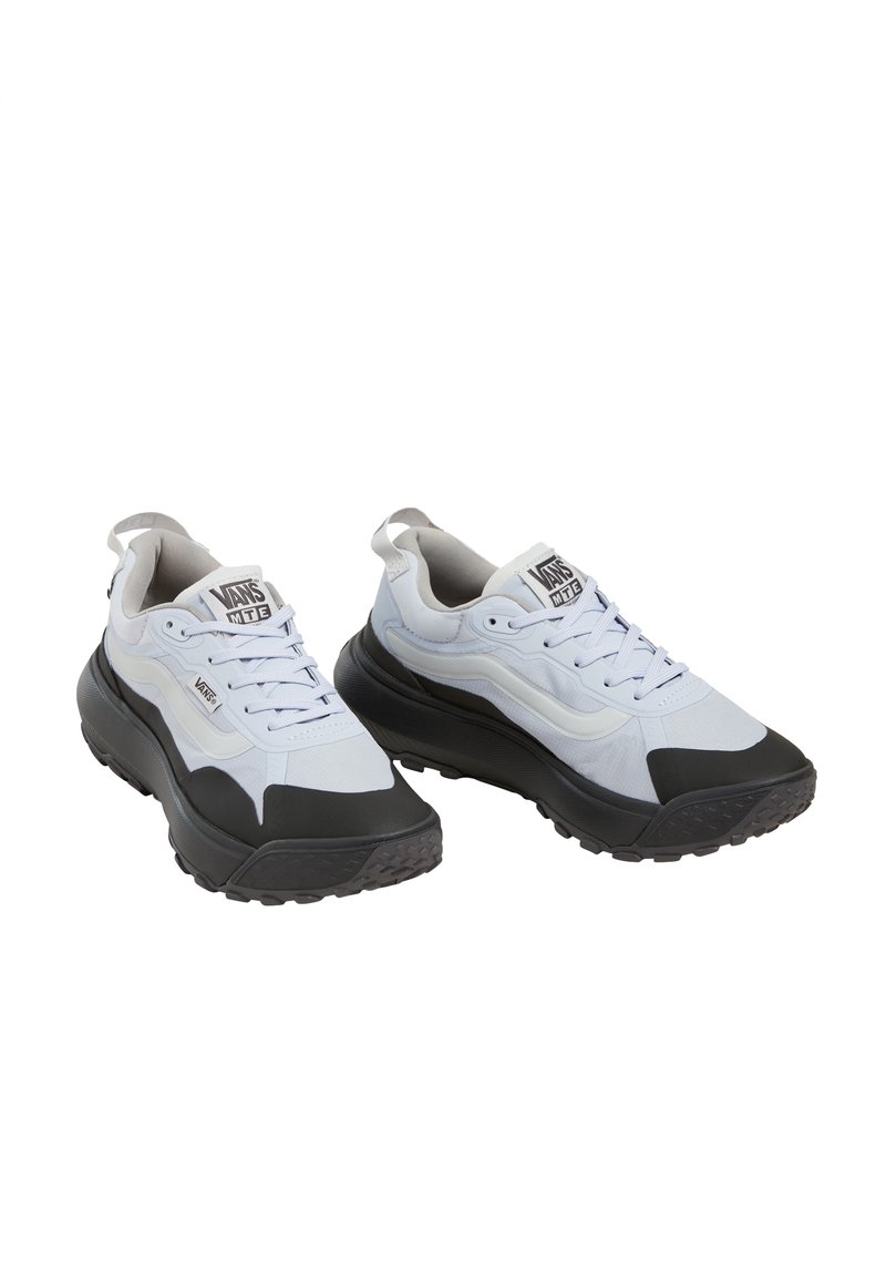 Un par de zapatillas Vans MTE en blanco y negro con suelas negras gruesas y cordones blancos, ligeramente inclinadas hacia la derecha sobre un fondo blanco.