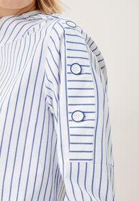 Vit skjorta med blå vertikala ränder, med en knappdetalj på ärmen och kontrasterande horisontella ränder nära manschetten.