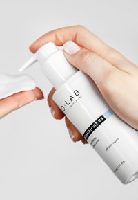 Biała cylindryczna butelka pompowa z etykietą "O LAB", zawierająca tonik z napisem "AHA + WIT B5", trzymana nad wacikiem. Czysty design, błyszczące wykończenie.