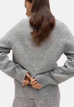 Grijze wollen trui met geribbelde manchetten en zoom. Oversized pasvorm met een getextureerd oppervlak. De handen van het model zijn achter de rug ineengeklemd, wat een ontspannen stijl toont.