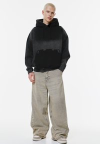 Felpa con gradiente nero e tasca a marsupio, abbinata a jeans grigi chiari a vestibilità ampia e sneakers beige, su uno sfondo bianco.