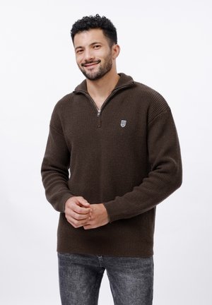 Brauner Reißverschluss-Pullover aus strukturiertem Strickstoff, mit einem kleinen Emblem auf der linken Brust. Kombiniert mit grauen Jeans.