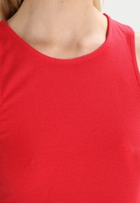 Haut rouge sans manches à côtes, avec un col arrondi, une coupe ajustée et une texture lisse avec une légère élasticité.