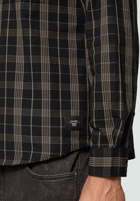 Chemise à carreaux noire et beige à manches longues, texture tissée, poche poitrine gauche et étiquette cousue près de l'ourlet.